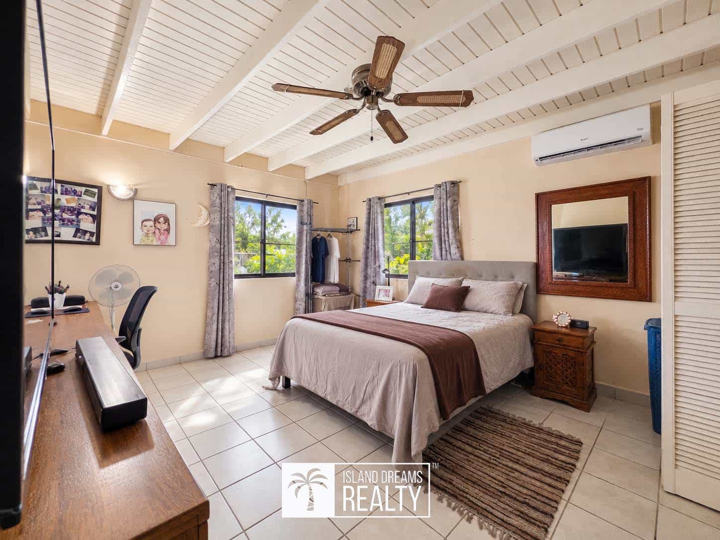 Bright master bedroom with a white beamed ceiling, ceiling fan, and a large bed dressed in beige and brown; two windows with patterned curtains flank the bed.