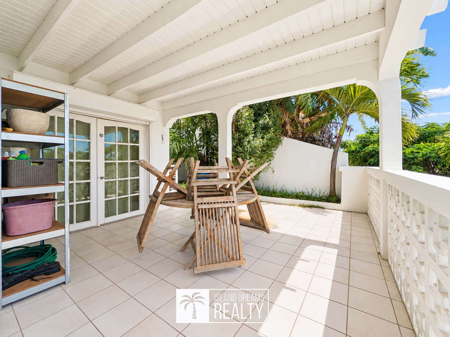 White covered patio with stacked wooden chairs on a tiled floor, glass French doors, and a tropical garden beyond.
