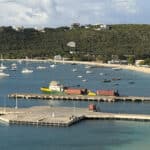 A marina with several sailboats anchored in calm blue water and a wooden pier extending into the bay, hillside houses in the background.