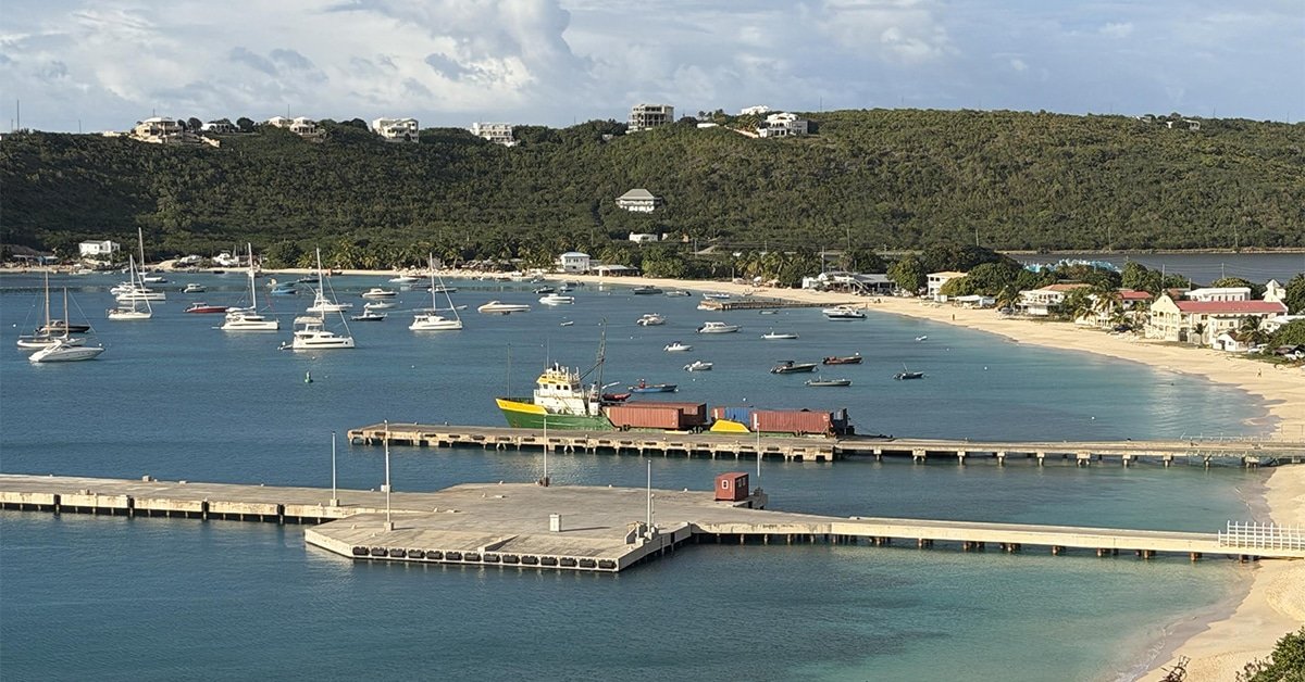 A marina with several sailboats anchored in calm blue water and a wooden pier extending into the bay, hillside houses in the background.