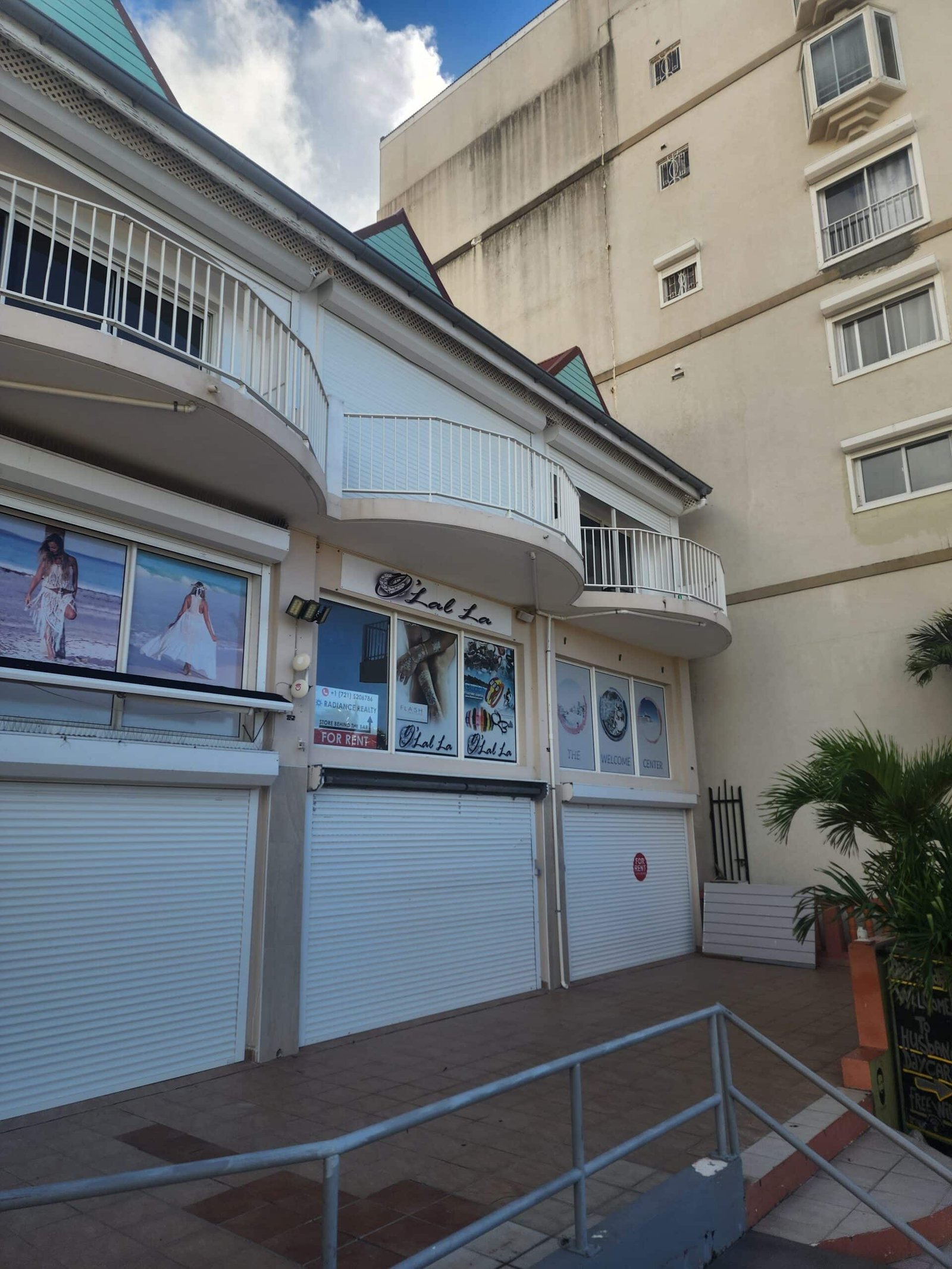 Shuttered storefront on a curved white balcony with railings, beige building behind, blue sky above neighboring structure with teal roof peaks.