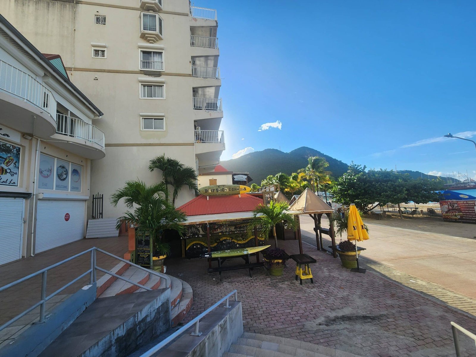 Tall beige building with balconies lining the left, stairs and a storefront at ground level, blue sky and distant mountains to the right; outdoor seating with yellow umbrellas nearby.