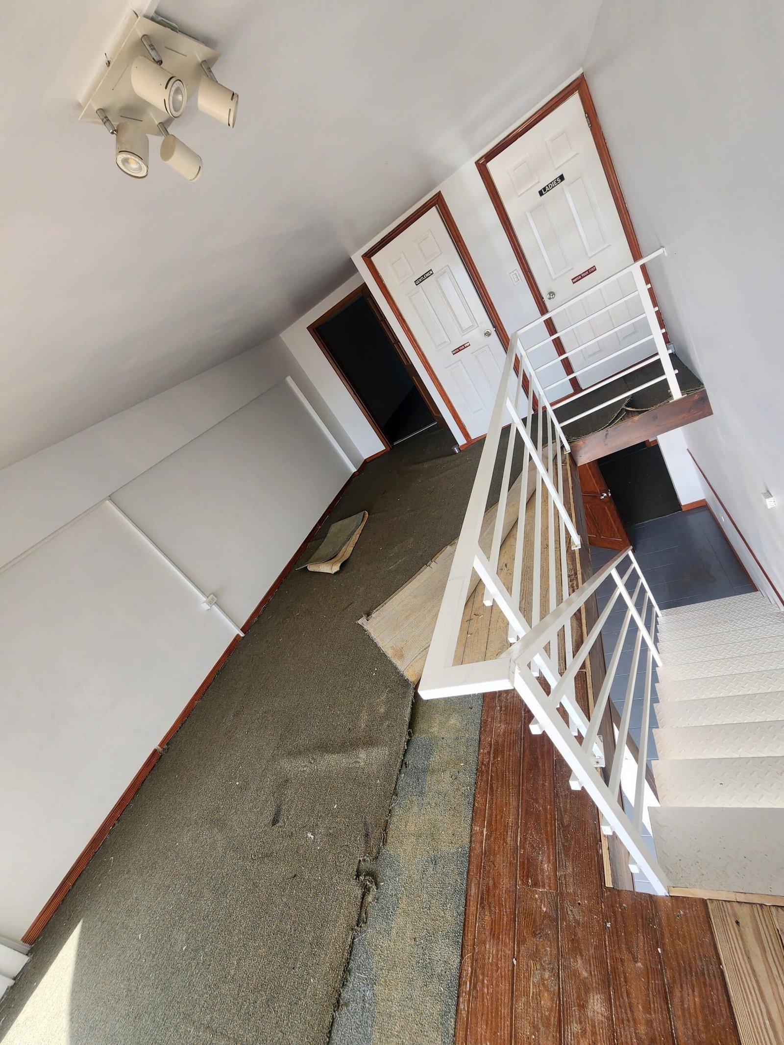Interior hallway with two white doors labeled Ladies and Gentlemen, white railing and wooden stairs nearby, carpeted floor and ceiling light fixture above.