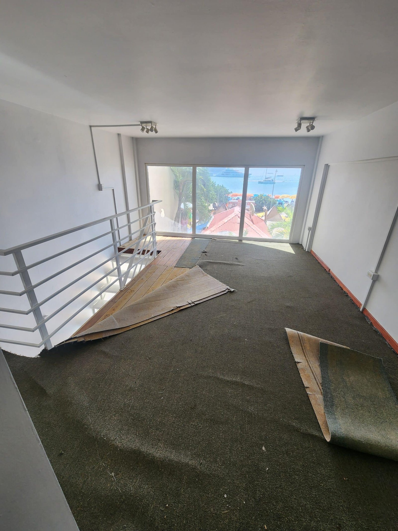 Interior stairwell with large window overlooking a beach and boats; damaged carpet and rolled rug lay on the floor near a metal railing.