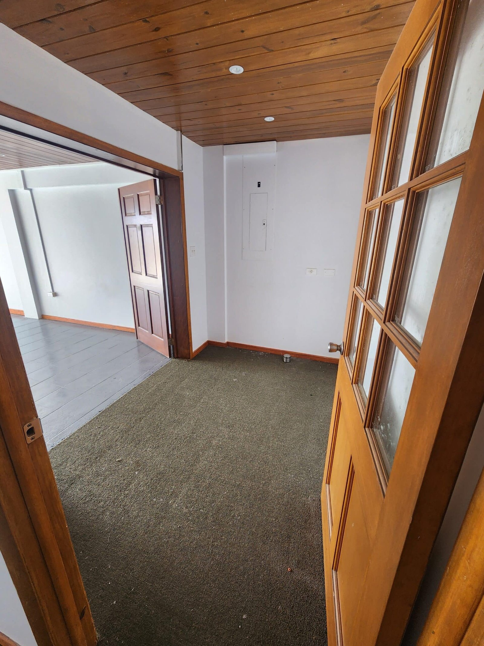 Open wooden door leading into a small entry area: wood-paneled ceiling, white walls, carpeted floor, and another wooden door visible to the left.