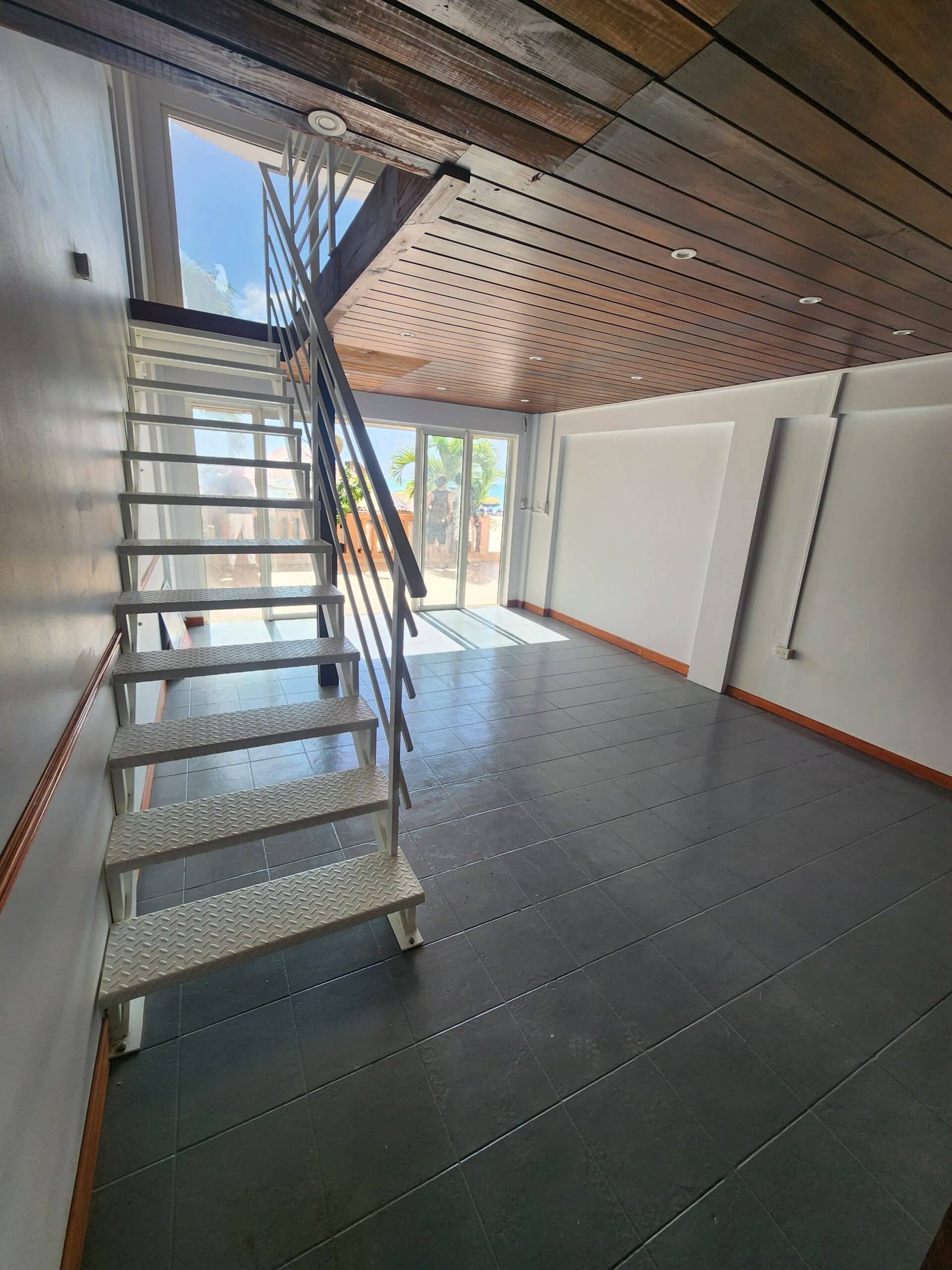 Industrial interior with metal staircase leading to a balcony opening to bright outdoors and palm trees visible through a large window.