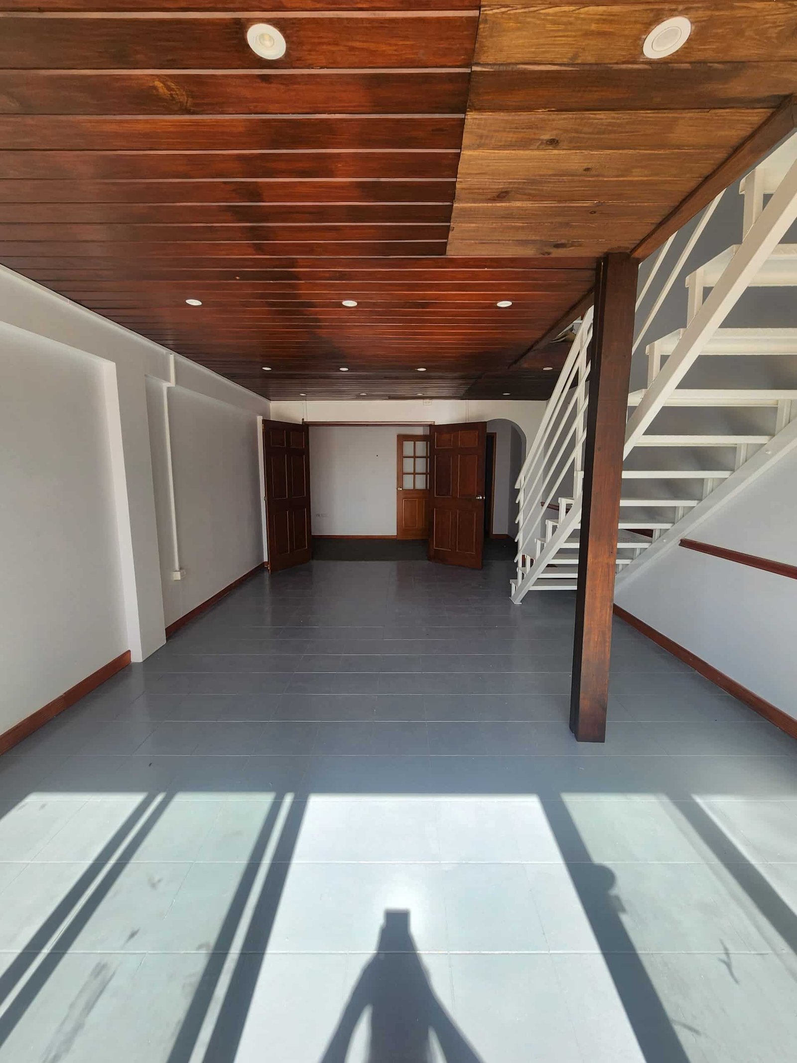 Interior of a spacious room with a dark wood plank ceiling, white staircase on the right, and double wooden doors open at the far end