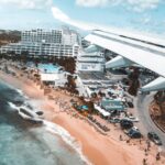 Aerial view of a beachfront resort with white hotel buildings, palm trees, a pool area, and a curved road along the shore; airplane wing in the foreground.