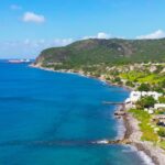Coastal village along a rocky shoreline with blue sea, white buildings, and green hills under a bright sky.