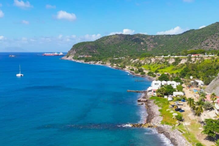 Coastal village along a rocky shoreline with blue sea, white buildings, and green hills under a bright sky.
