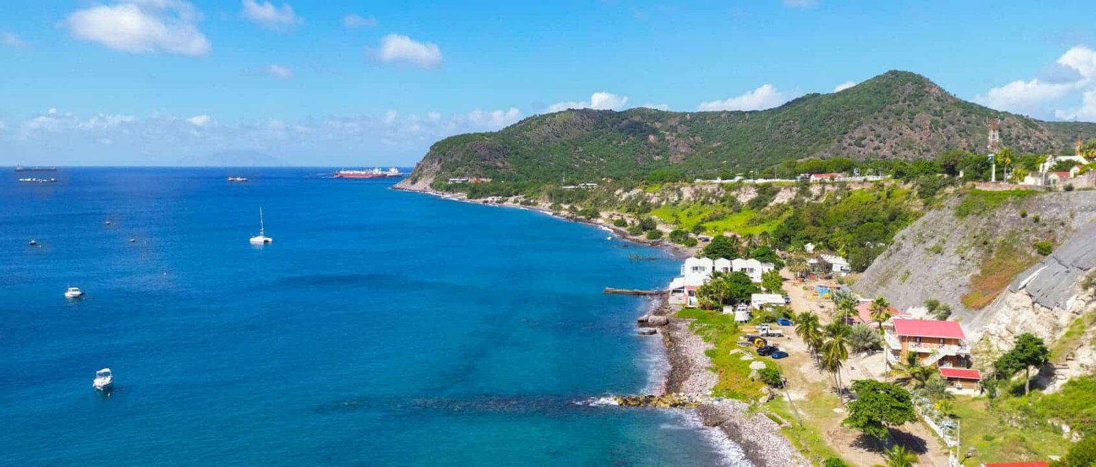 Coastal village along a rocky shoreline with blue sea, white buildings, and green hills under a bright sky.
