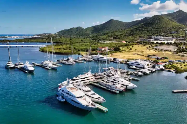 A marina filled with white yachts docked along piers, turquoise water and green hills in the background.