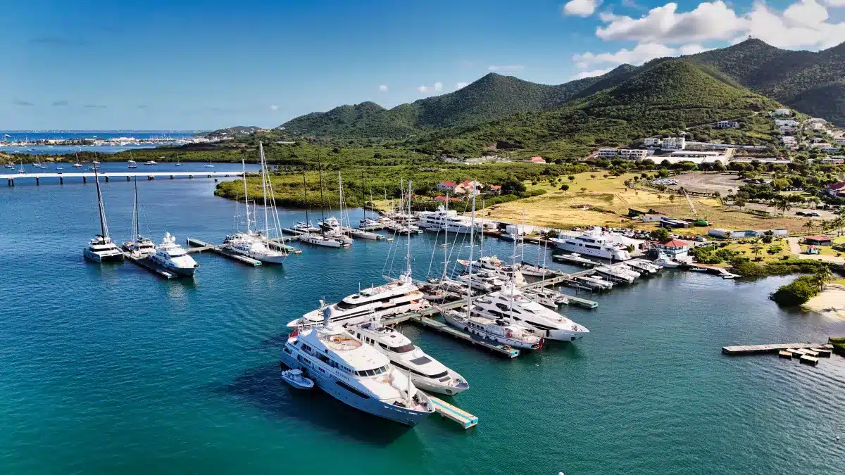 A marina filled with white yachts docked along piers, turquoise water and green hills in the background.