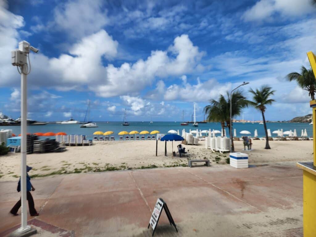 Soleil Commercial Complex Unit 61 IDR Tropical beach scene with yellow umbrellas, white lounge chairs, palm trees, and boats on calm blue water under a bright sky; promenade in the foreground.