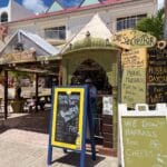 Open-air tropical bar scene with carved green kiosk and chalkboard menus advertising cocktails and snacks. Mom-and-pop beach vibe.