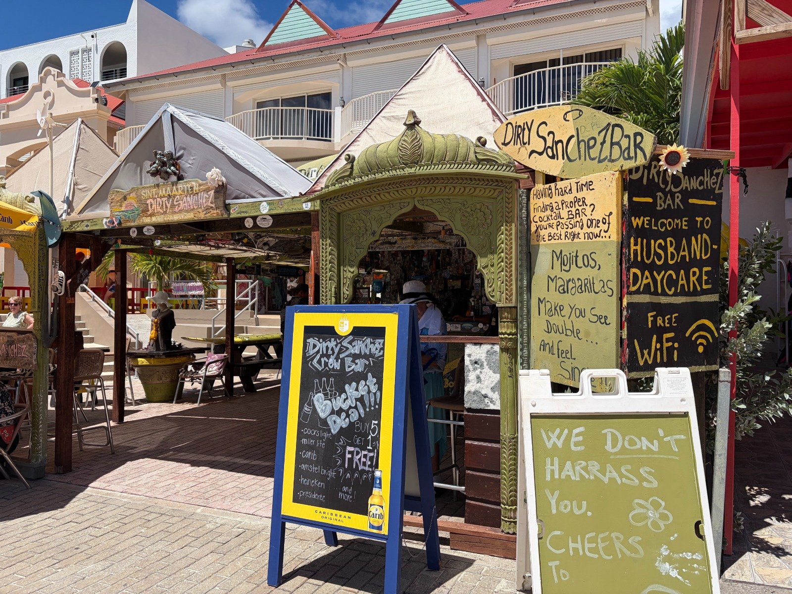 Open-air tropical bar scene with carved green kiosk and chalkboard menus advertising cocktails and snacks. Mom-and-pop beach vibe.