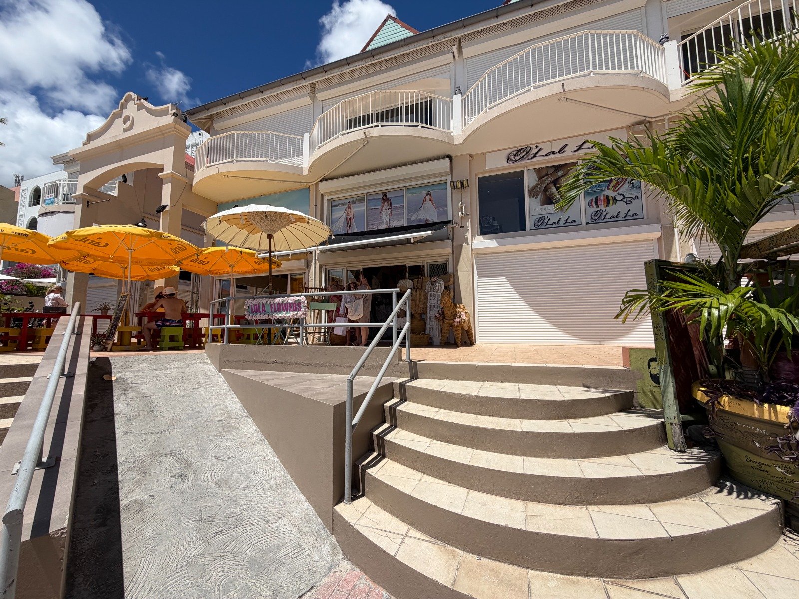 Outdoor shopping street with yellow parasols, seating, and a clothing shop under a two-story beige building with balconies.