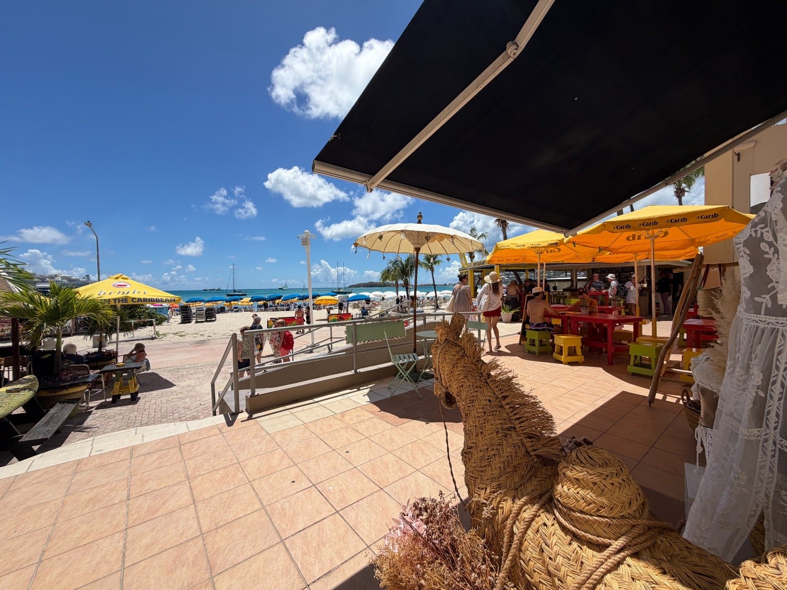 Beachfront cafe with yellow umbrellas and people seated at colorful tables overlooking a sandy beach and blue sea under a sunny sky.