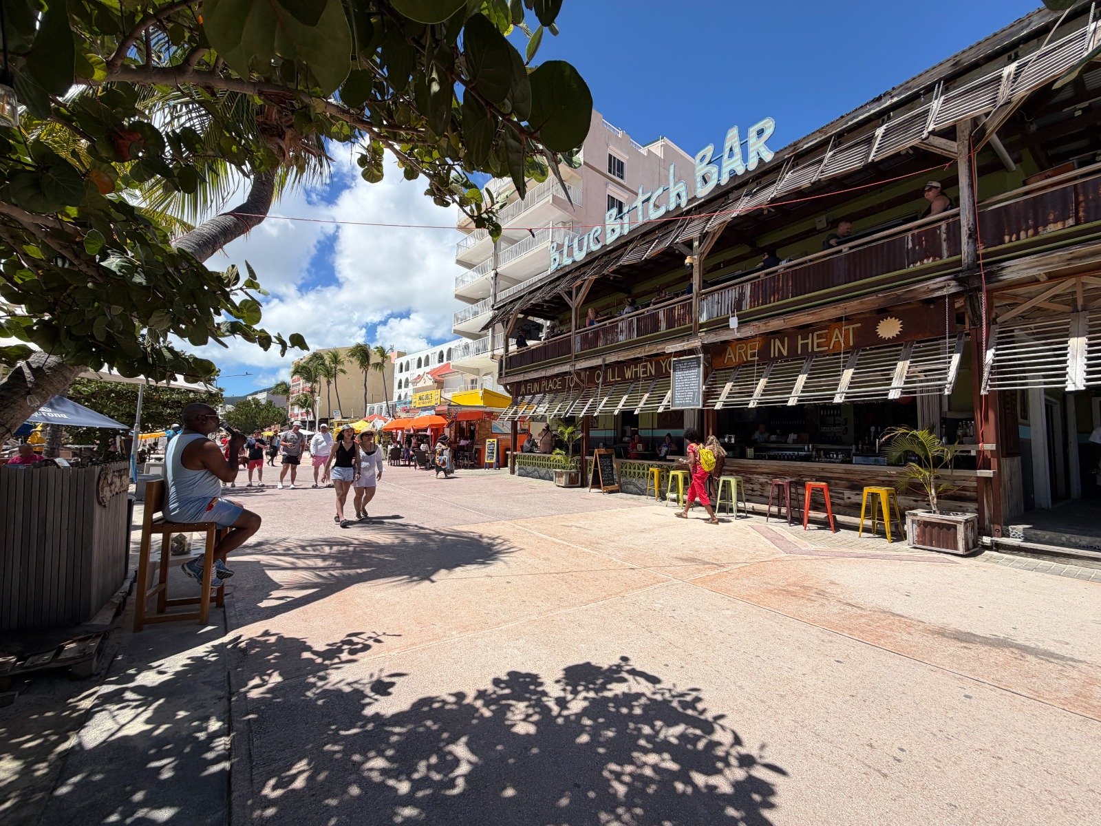 Sunlit street scene with a two-story rustic bar, open-air seating, and pedestrians walking past under palm trees.