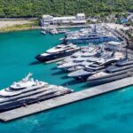 Marina crowded with luxury yachts docked along concrete piers on turquoise water, with a tree-lined shore in the background.