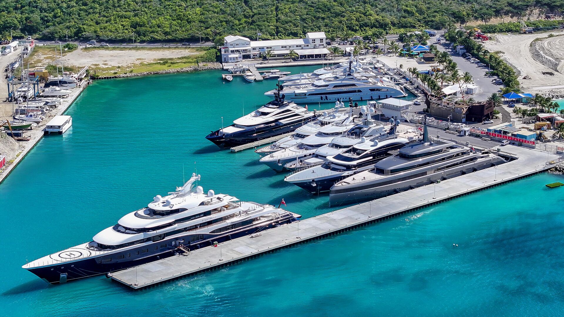 Marina crowded with luxury yachts docked along concrete piers on turquoise water, with a tree-lined shore in the background.