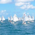 Group of small sailboats with white sails racing on blue water near a rocky jetty, sunny sky with a few clouds, and palm trees in the background.