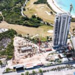 Aerial view of a construction site beside a tall residential tower near a blue coastline and beach, with cranes and cleared land nearby
