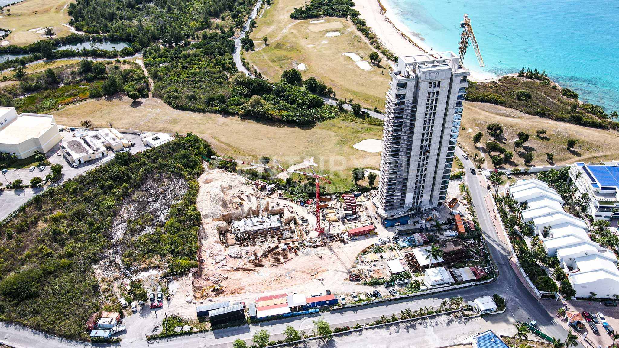 Aerial view of a construction site beside a tall residential tower near a blue coastline and beach, with cranes and cleared land nearby