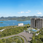 Aerial view of a coastal resort with tall white apartment buildings, a large pool, palm trees, and a bay with islands and green mountains in the distance.