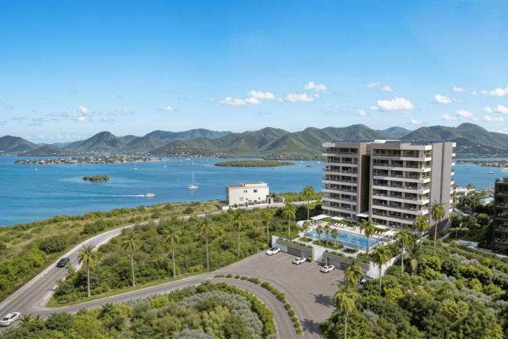 Aerial view of a coastal resort with tall white apartment buildings, a large pool, palm trees, and a bay with islands and green mountains in the distance.