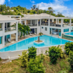 Aerial view of a modern white villa with a large blue swimming pool and a small island palm tree in the center courtyard.