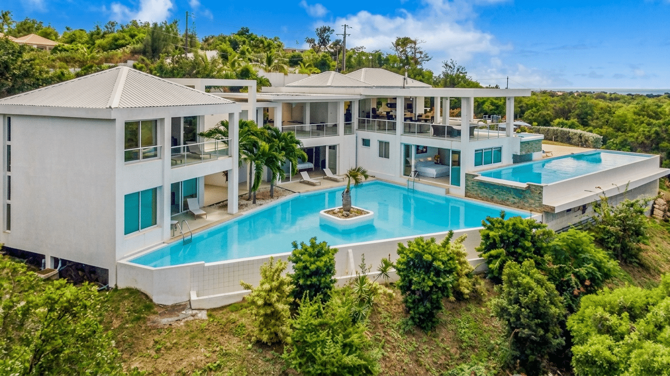 Aerial view of a modern white villa with a large blue swimming pool and a small island palm tree in the center courtyard.