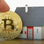 Close-up of a gold Bitcoin coin being held beside a miniature gray-and-red house on a wooden surface.
