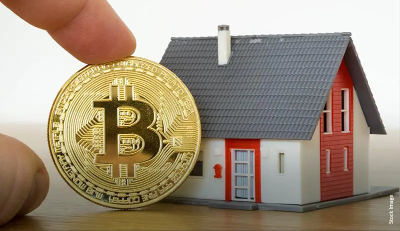 Close-up of a gold Bitcoin coin being held beside a miniature gray-and-red house on a wooden surface.