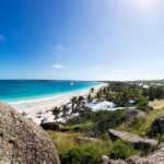 Panoramic view of a tropical beach with turquoise water, white sand, palm trees, and rocky cliffs in the foreground on a sunny day.