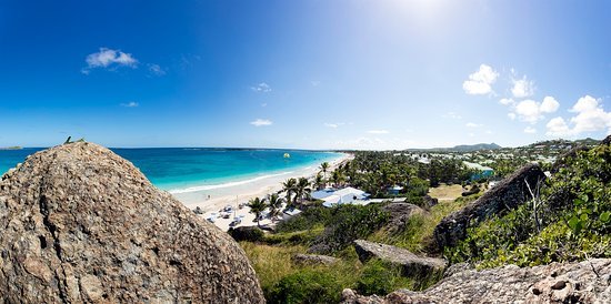 Panoramic view of a tropical beach with turquoise water, white sand, palm trees, and rocky cliffs in the foreground on a sunny day.