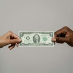 Two hands pass a US two-dollar bill to each other against a gray background.
