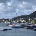 Harbor scene with several sailboats docked at a marina, calm water in the foreground and a town with hills in the background.