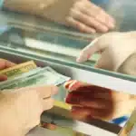 Person hands full of cash being passed through an ATM or bank counter window, indicating a cash transaction