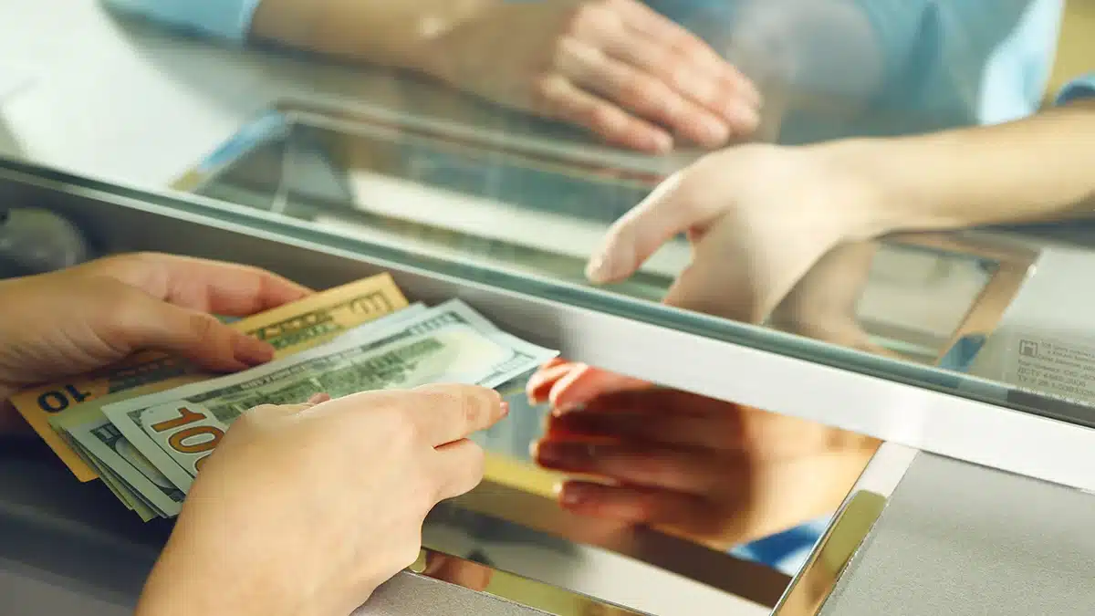 Person hands full of cash being passed through an ATM or bank counter window, indicating a cash transaction