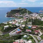 A seaside hillside town with winding roads and colorful houses overlooking the ocean; a cruise ship is docked at the harbor.
