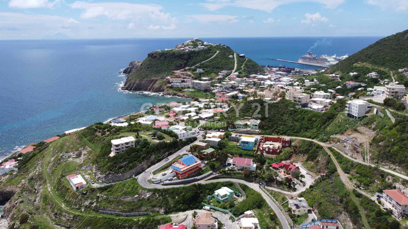 A seaside hillside town with winding roads and colorful houses overlooking the ocean; a cruise ship is docked at the harbor.