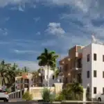 Modern white residential buildings with balconies and palm trees along a gated street at dusk, string lights overhead, several parked cars nearby