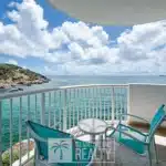 Balcony seating area with two turquoise chairs and a small glass table overlooking clear blue water and a rocky coast under a sunny, partly cloudy sky.