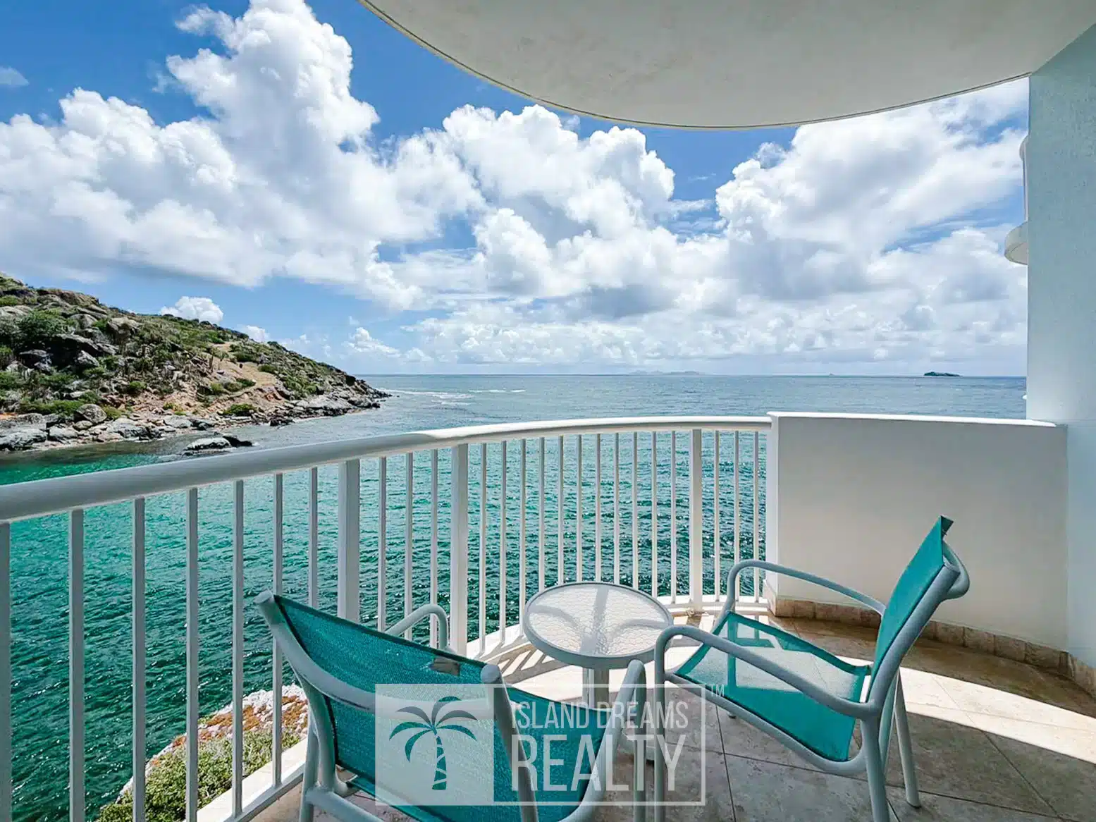 Balcony seating area with two turquoise chairs and a small glass table overlooking clear blue water and a rocky coast under a sunny, partly cloudy sky.