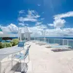 Rooftop terrace with white and blue lounge chairs and round tables, overlooking the ocean under a bright blue sky.