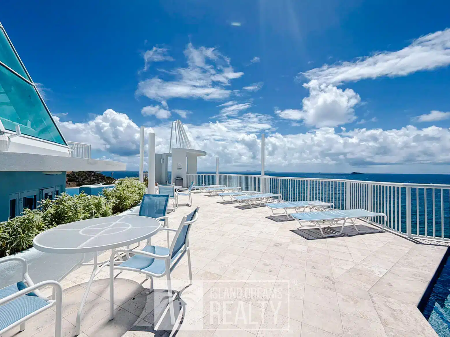 Rooftop terrace with white and blue lounge chairs and round tables, overlooking the ocean under a bright blue sky.