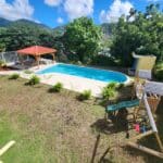 Backyard with a kidney-shaped pool, red-roofed gazebo, and wooden play structures against green hills and blue sky.