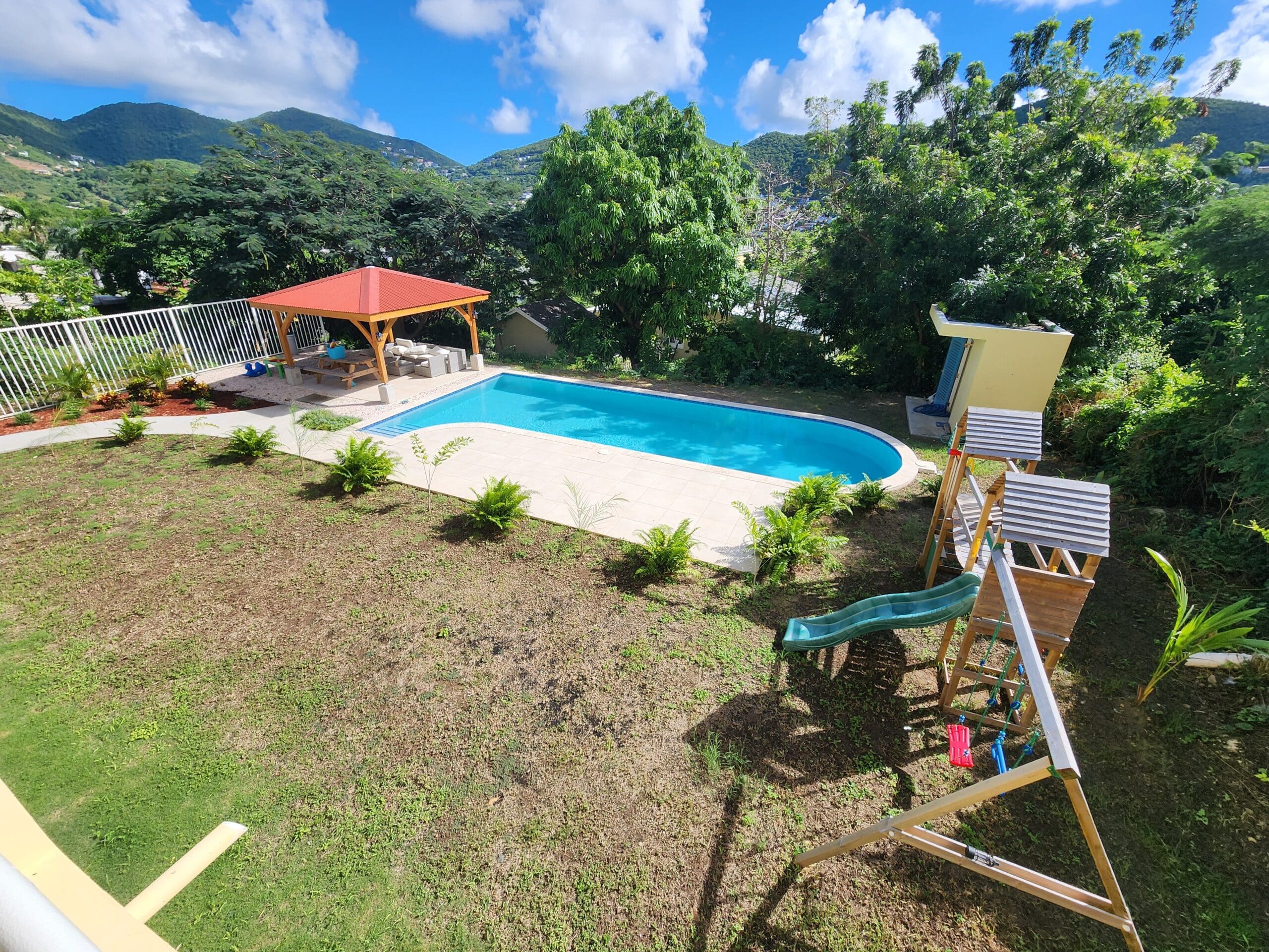 Backyard with a kidney-shaped pool, red-roofed gazebo, and wooden play structures against green hills and blue sky.