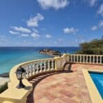Terrace overlooking the blue sea with a yellow balustrade and terracotta tiles, a pool on the right, and a sunny sky above.